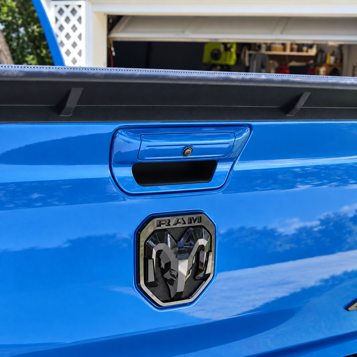 Close-up of the rear tailgate of a bright blue Ram truck, showing the black tailgate handle and a gloss black dual-intensity LED Ram tailgate logo badge on the tailgate