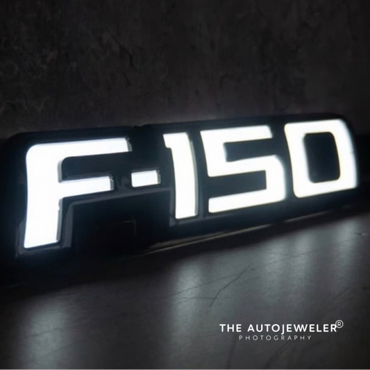 Illuminated gloss black F-150 LED fender emblem glowing white in a dark setting, angled close-up view.