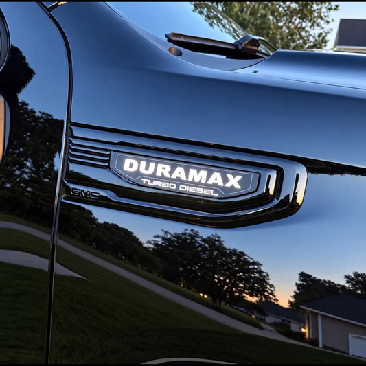 Close-up of a glossy blue GMC Duramax Turbo Diesel truck body panel with the chrome badge visible, reflecting a suburban outdoor scene at dusk.