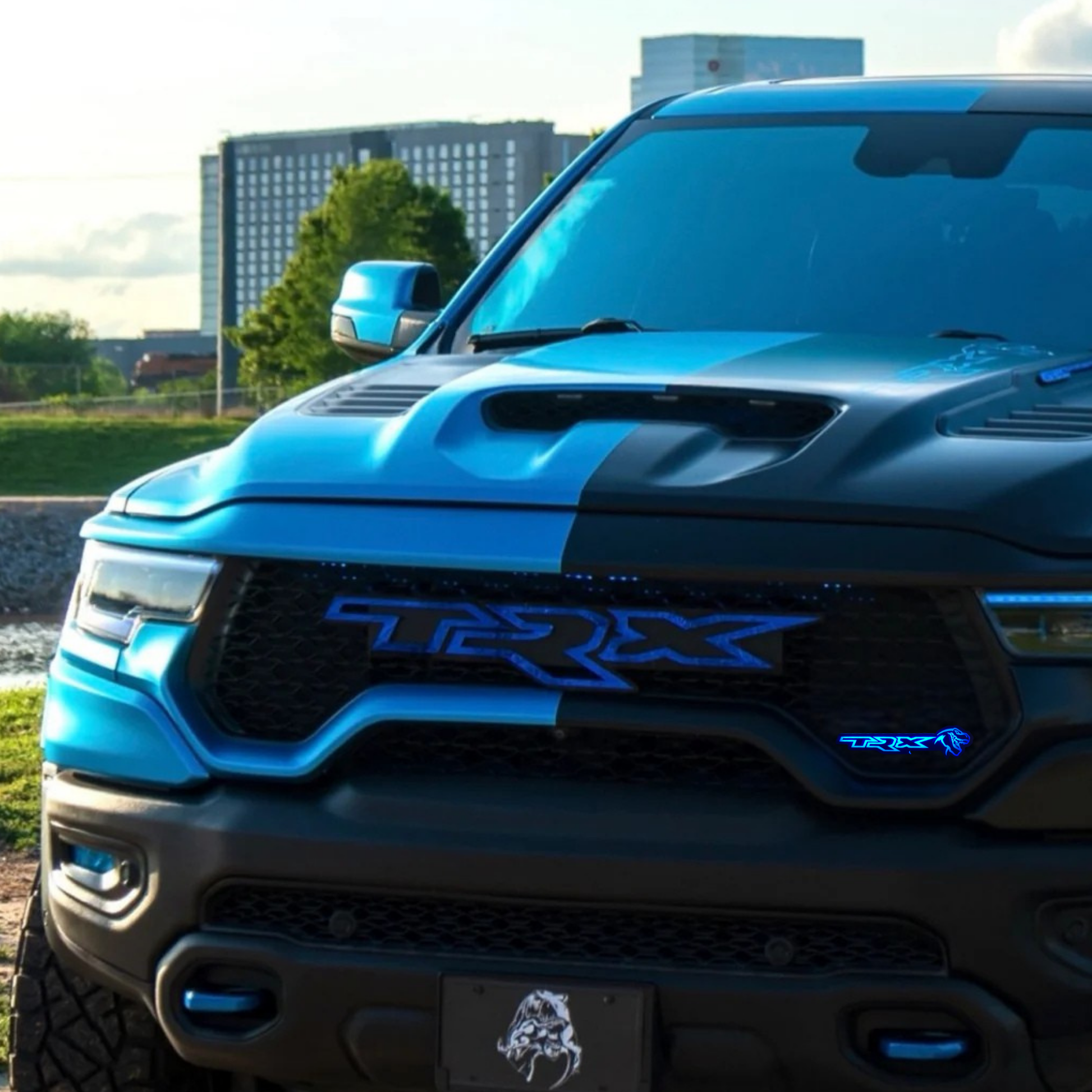 Blue and black RAM TRX truck with blue LED grille emblem, parked in front of buildings in an urban setting.