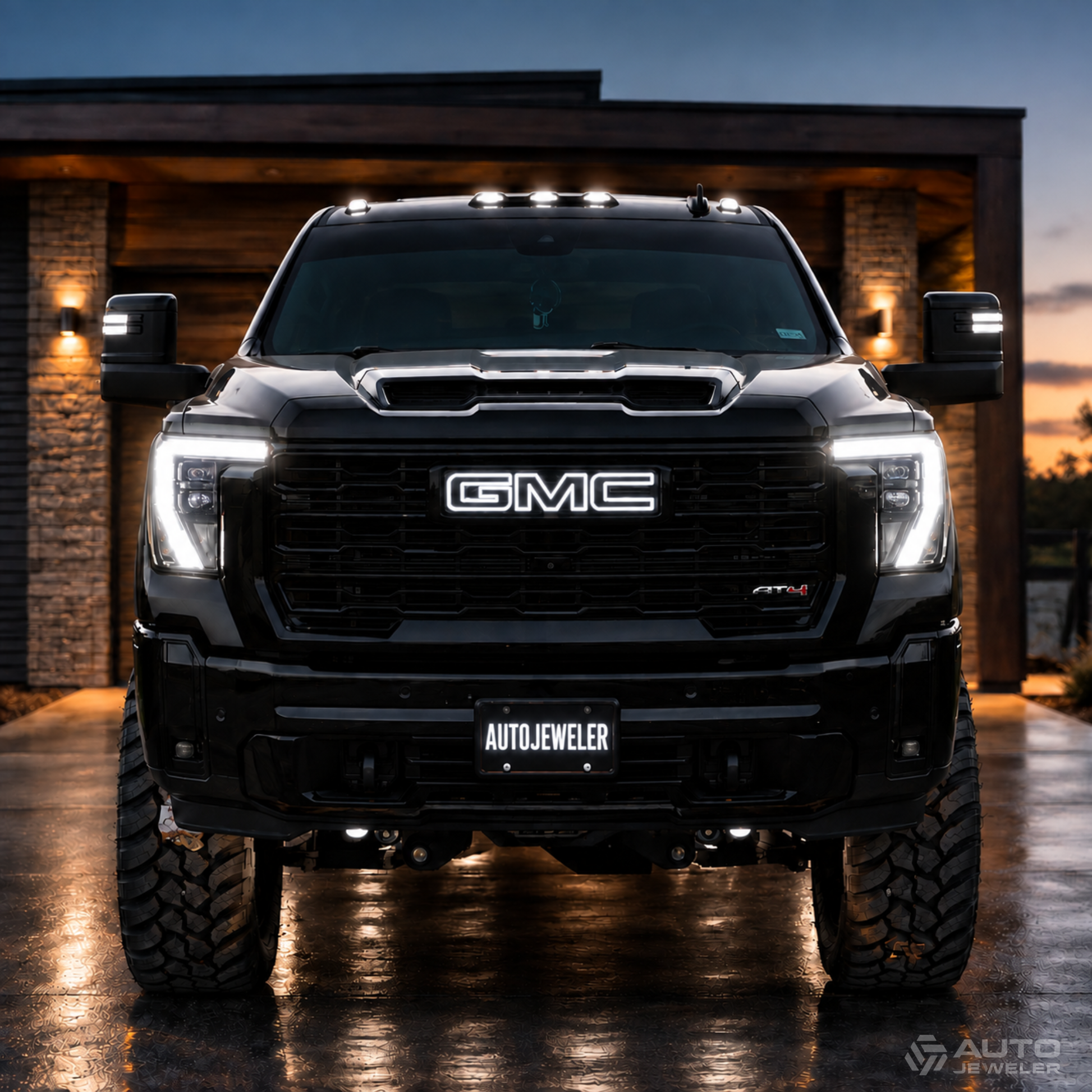 A black GMC pickup truck photographed head-on in a driveway at dusk, with its white headlights and side marker lights turned on. The truck grille prominently shows the white illuminated GMC logo, and an AUTOJEWELER LED license plate is mounted on the front lower bumper.