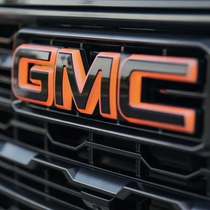A close-up shot of a glossy black LED illuminated GMC emblem with a vibrant amber glow, mounted on a black GMC truck grille.