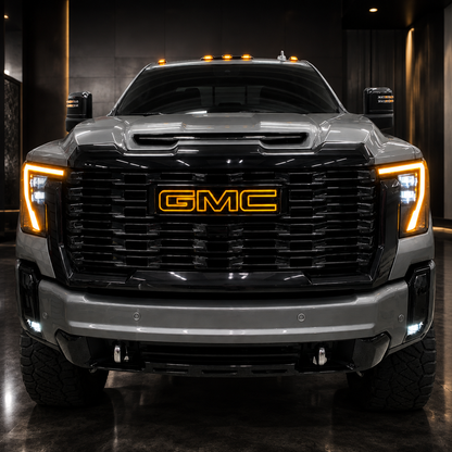 A front-facing view of a large silver and black GMC pickup truck with illuminated amber LED accents around the grille, an AutoJeweler LED GMC grille logo illuminating in amber, and headlights, photographed in a dark indoor showroom or studio setting.