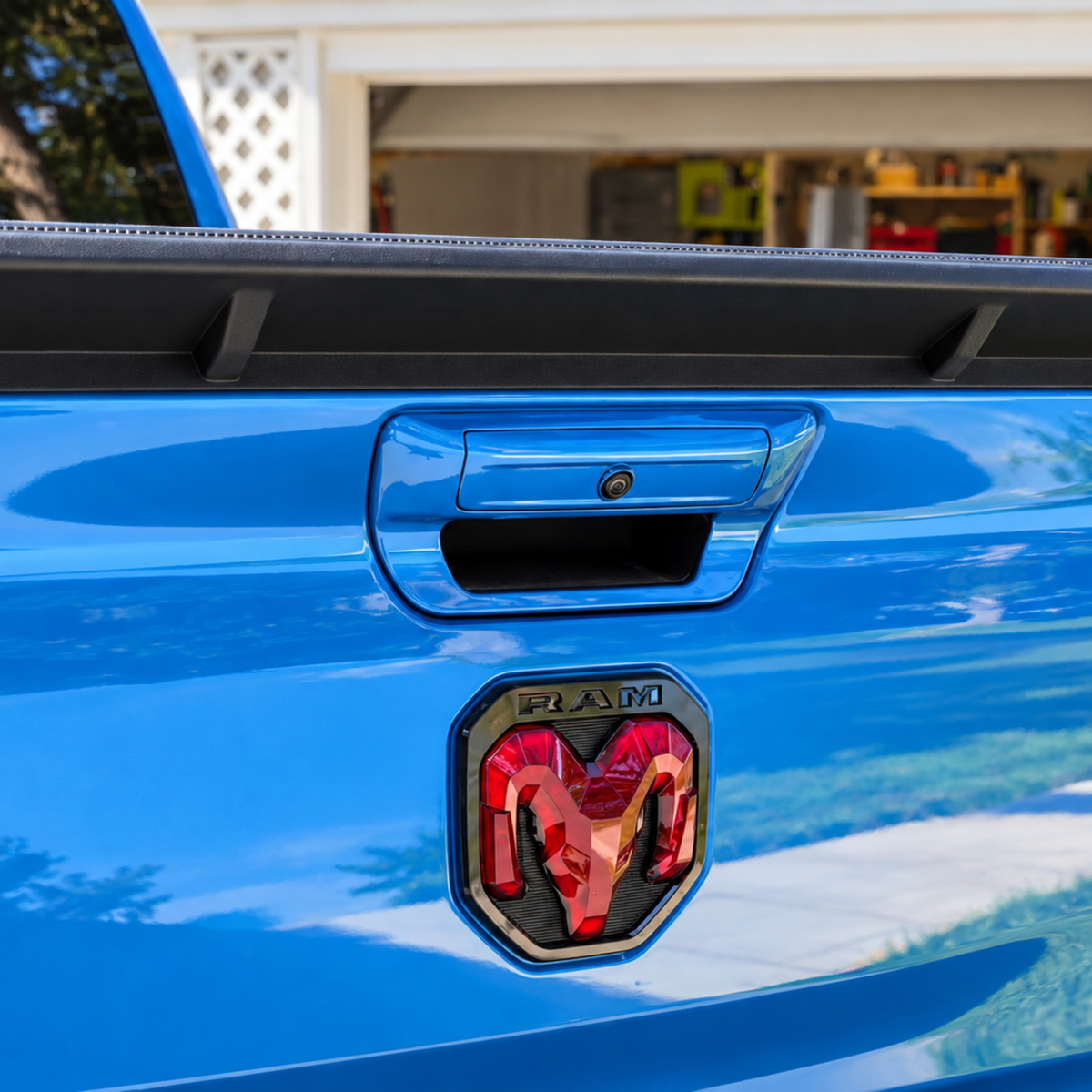 Close-up of the rear tailgate of a bright blue Ram truck, showing the black tailgate handle, a black spoiler edge, and the red LED Illuminated  black Ram logo badge.