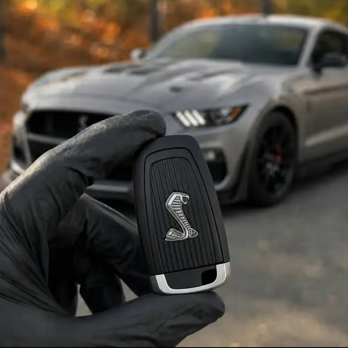 A hand wearing a black glove holds a black and silver Shelby key fob in the foreground, with a blurred gray Mustang Shelby car