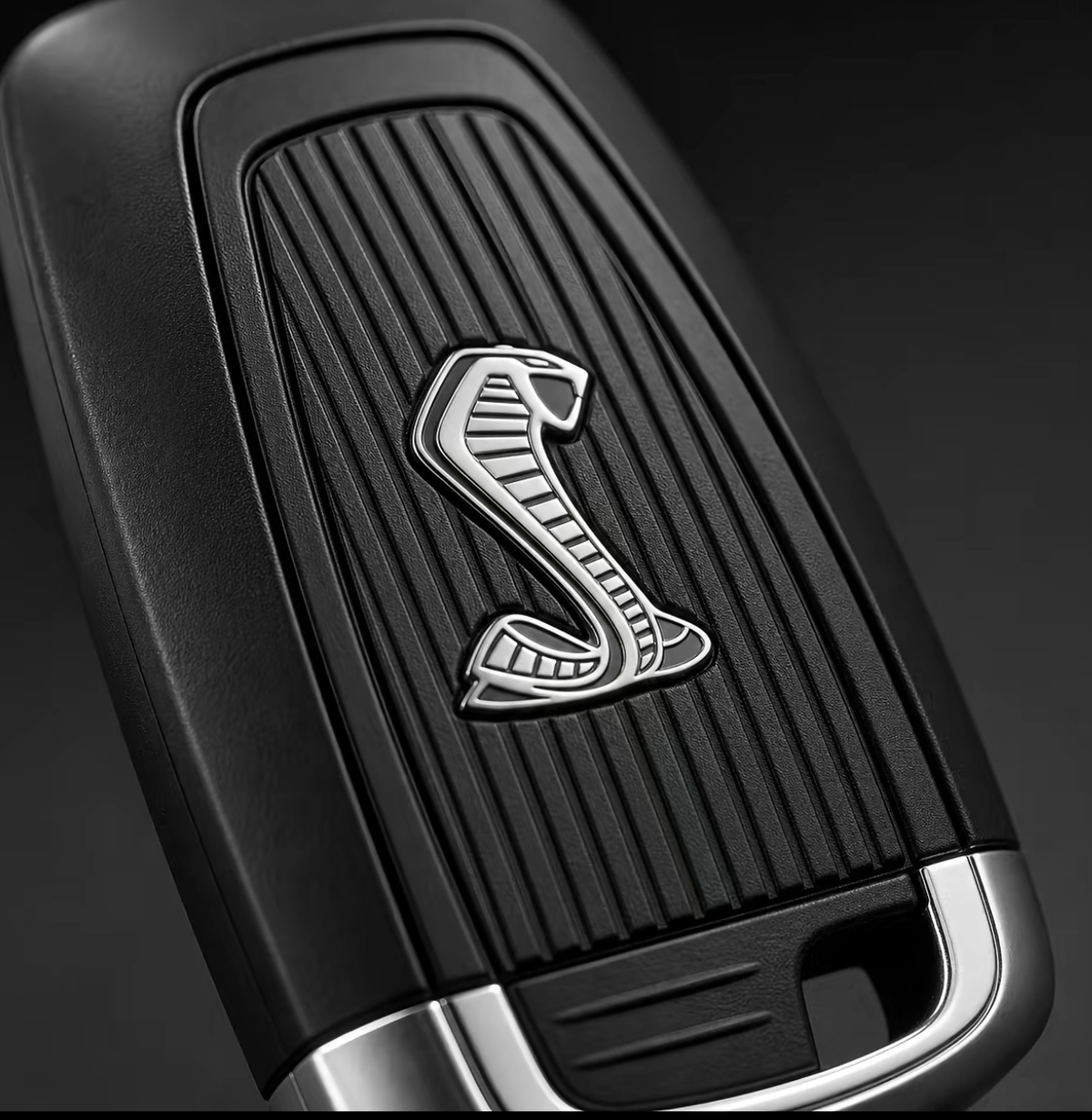 Close-up of a black car key fob featuring a silver Cobra logo on a ribbed textured panel, with a polished silver metal accent at the bottom, set against a dark background.