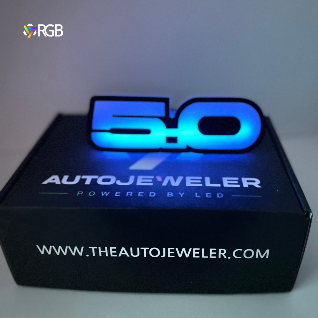 RGB LED Flow-Series 5.0 Logo Emblem glowing blue on top of an AutoJeweler box.