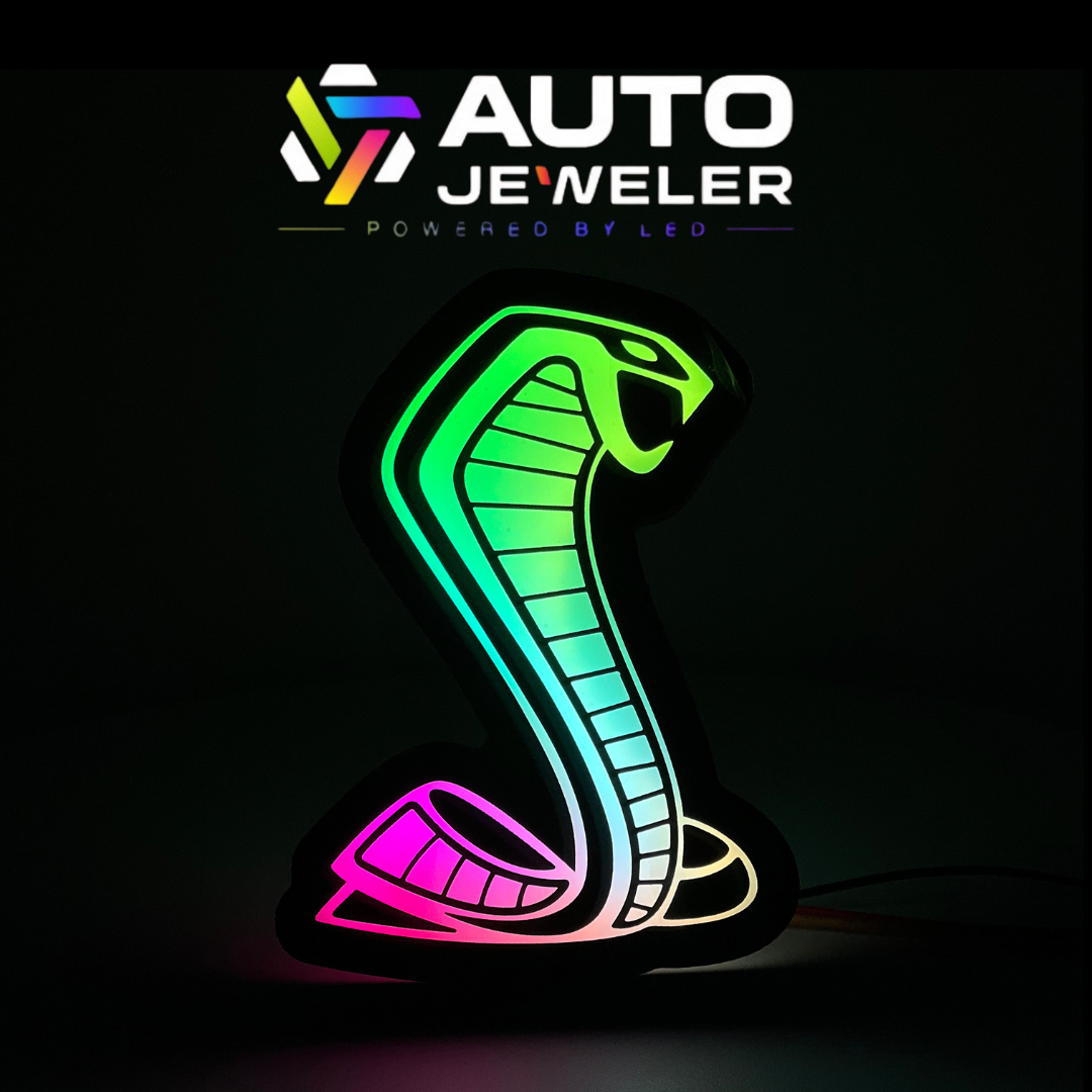 AutoJeweler RGBW Mustang Cobra grille emblem illuminated with full-spectrum flow lighting, displayed under branded banner background