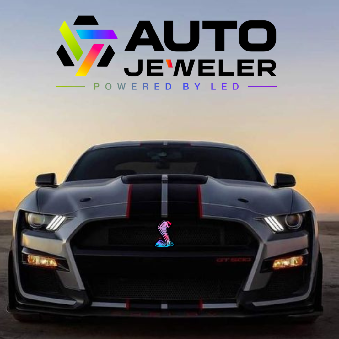  Dark gray Ford Mustang Shelby GT500 at sunset featuring an AutoJeweler RGBW light-up Cobra grille emblem glowing in blue and pink, with AutoJeweler logo above the car