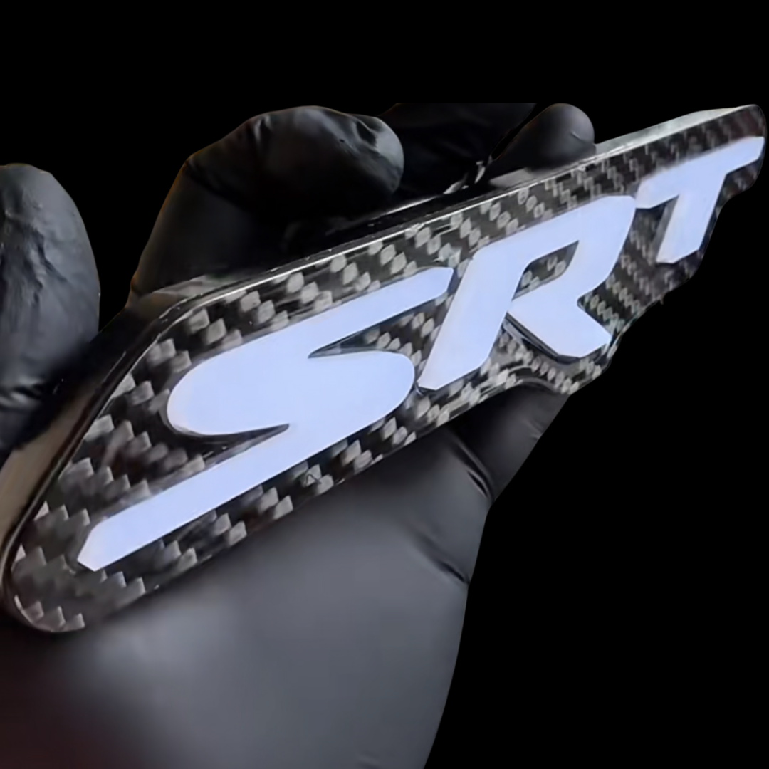 Angled close-up of the carbon fiber SRT LED emblem powered off with white raised lettering held in a black glove.