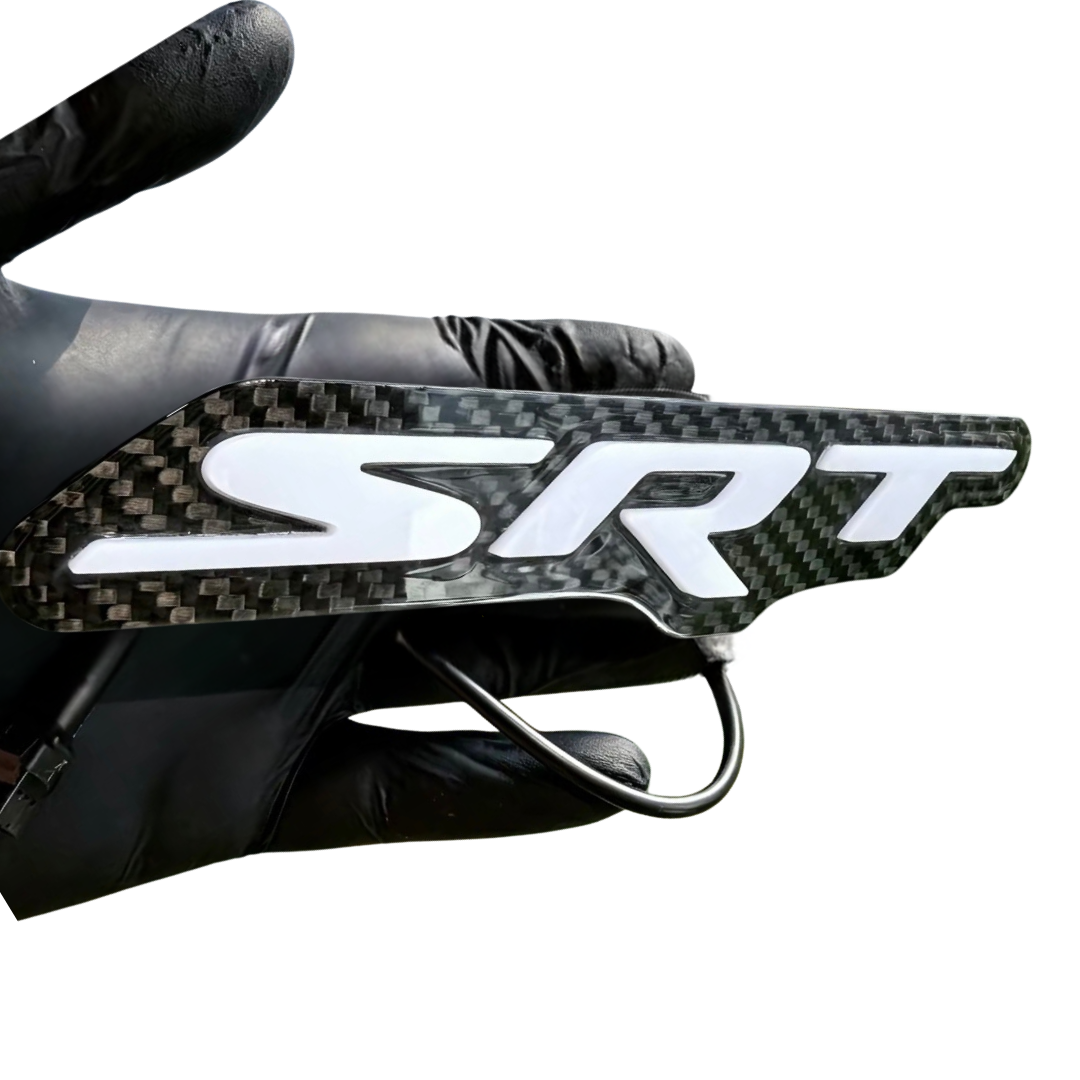 Close-up of an AutoJeweler carbon fiber SRT LED grille emblem held in a black glove with white raised lettering.