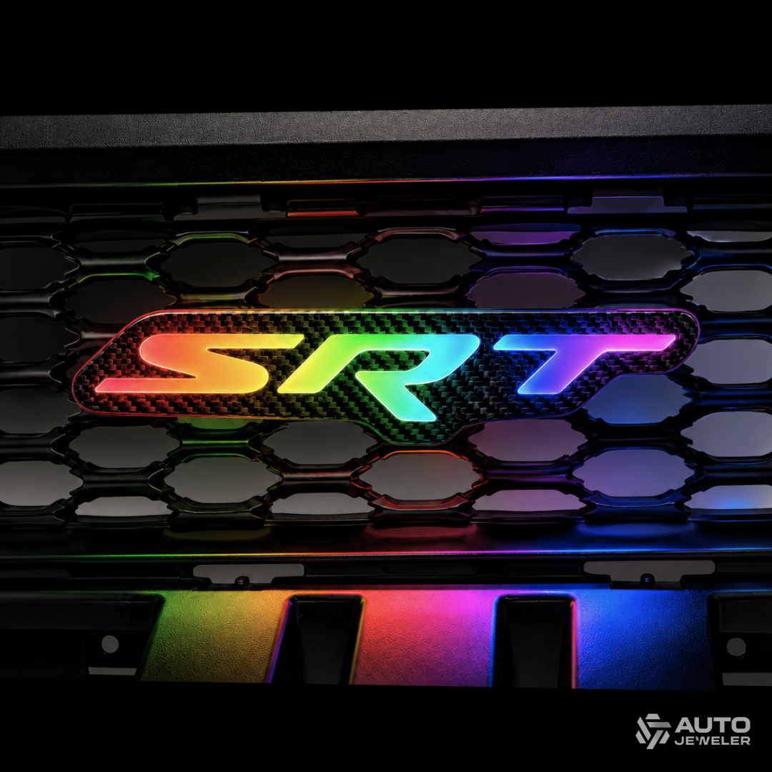 Close-up of an AutoJeweler carbon fiber SRT RGB Flow Series grille emblem glowing in a vivid rainbow gradient.