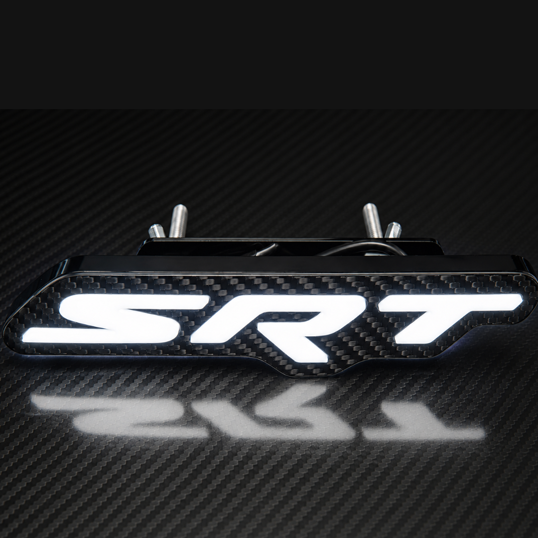 A carbon fiber textured SRT emblem badge with bright white illuminated lettering, shown on a dark carbon fiber surface with a reflection below.