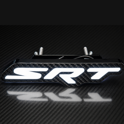 A carbon fiber textured SRT emblem badge with bright white illuminated lettering, shown on a dark carbon fiber surface with a reflection below.