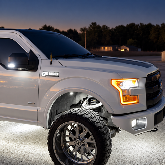Illuminated white F-150 fender badge on a lifted silver-white truck at dusk, beside amber headlights and white wheel-well lighting.