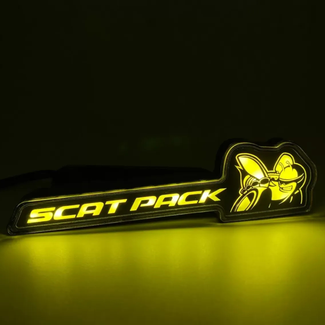Scat Pack RGB Flow-Series LED grille emblem glowing yellow on a black background – AutoJeweler product close-up.