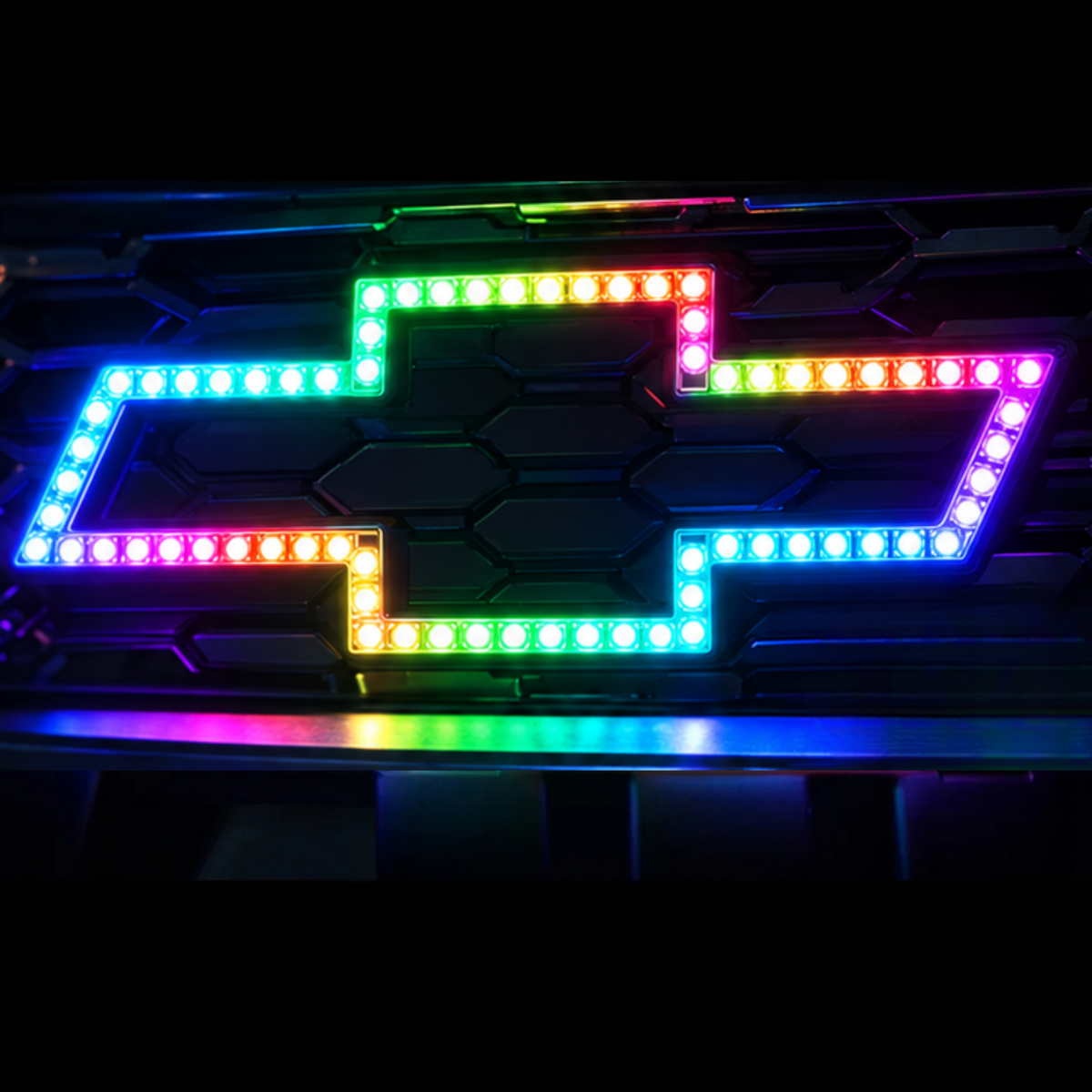 Close-up of the AutoJeweler Chevy Flow Series LED bowtie emblem illuminated in a multicolor pattern on a black grille.