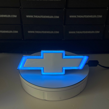 Chevy Silverado RGB LED Bowtie Grille Emblem illuminated in blue light on AutoJeweler display stand.