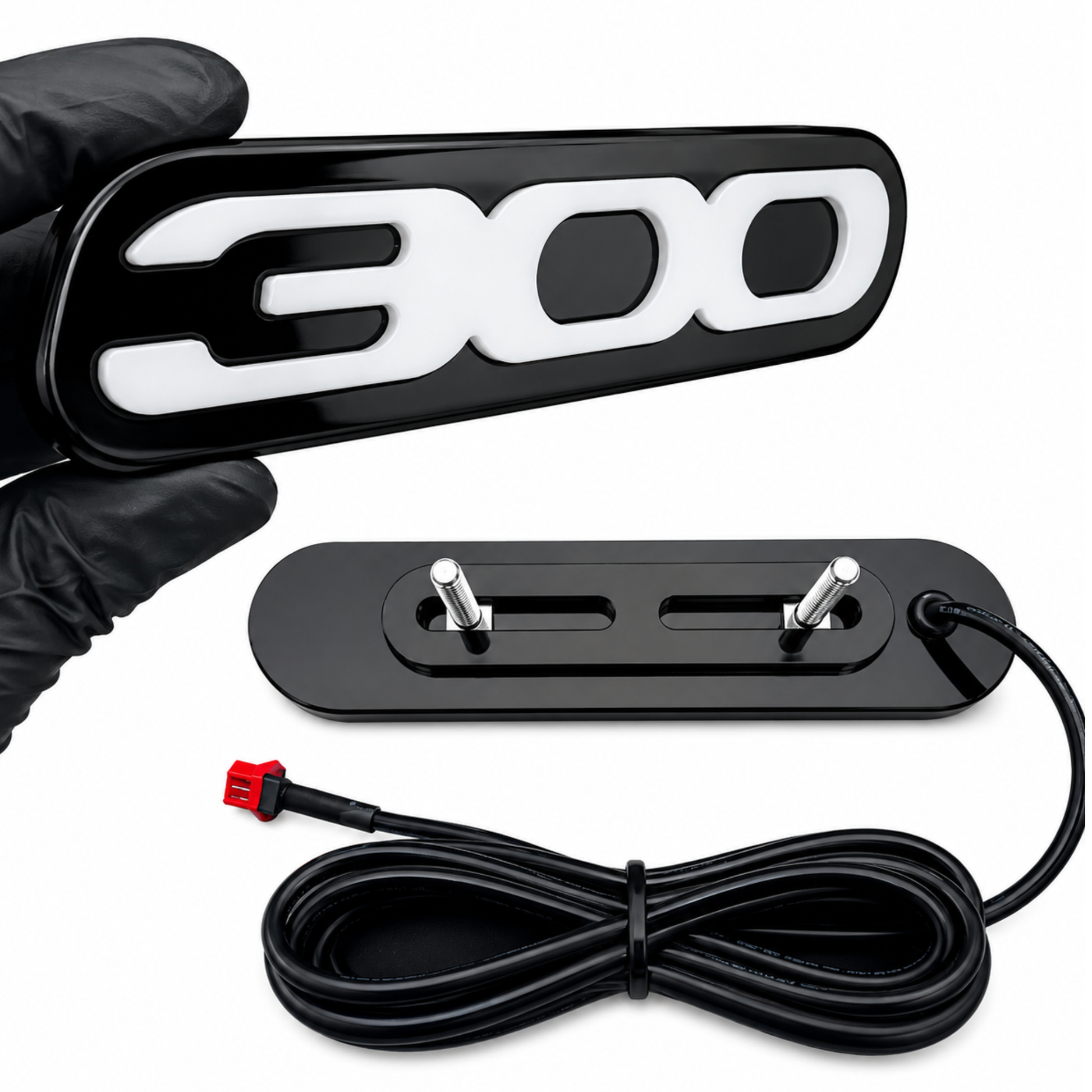 Front and rear view of Chrysler 300 LED grille emblem showing gloss black acrylic faceplate, adjustable mounting bracket, threaded studs, and plug-and-play wiring harness.