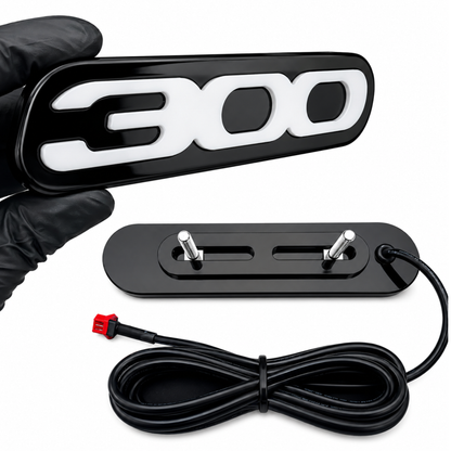 Front and rear view of Chrysler 300 LED grille emblem showing gloss black acrylic faceplate, adjustable mounting bracket, threaded studs, and plug-and-play wiring harness.