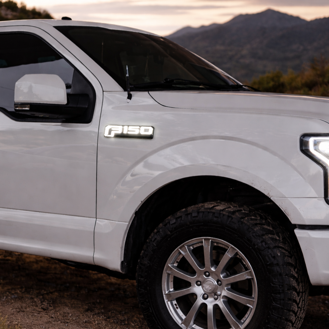AutoJeweler LED F-150 lighted fender emblem installed on white Ford F-150 truck outdoors at sunset showing illuminated badge in lifestyle setting.