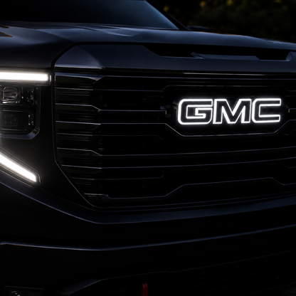 Close-up of a black GMC truck front grille with illuminated white GMC lettering and bright LED headlights in a dark outdoor setting.