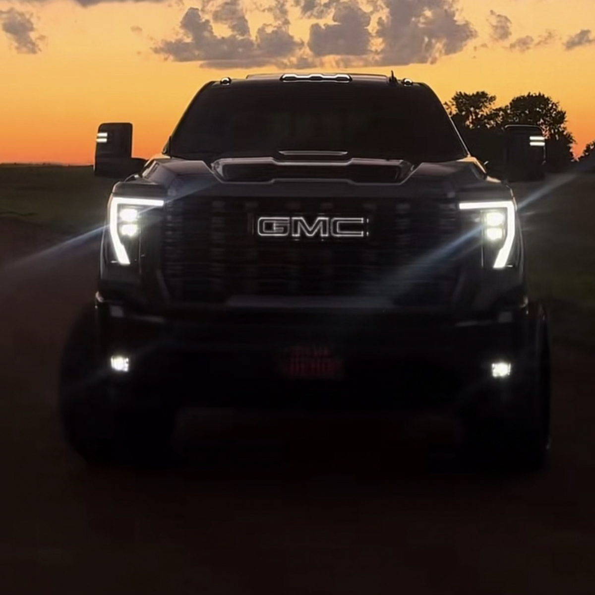 A black GMC pickup truck photographed head-on on a dirt road at sunset, with the GMC grille and logo clearly visible illuminated in a bright white color and the vehicle lights turned on.