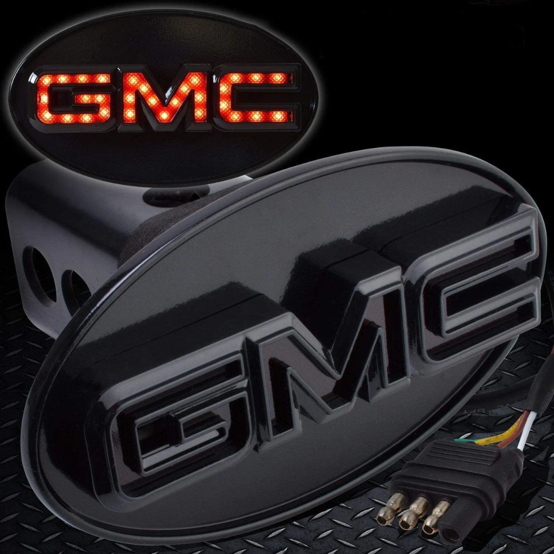 Close-up gloss black GMC LED hitch cover showing red illuminated lettering and 4-way trailer plug connector