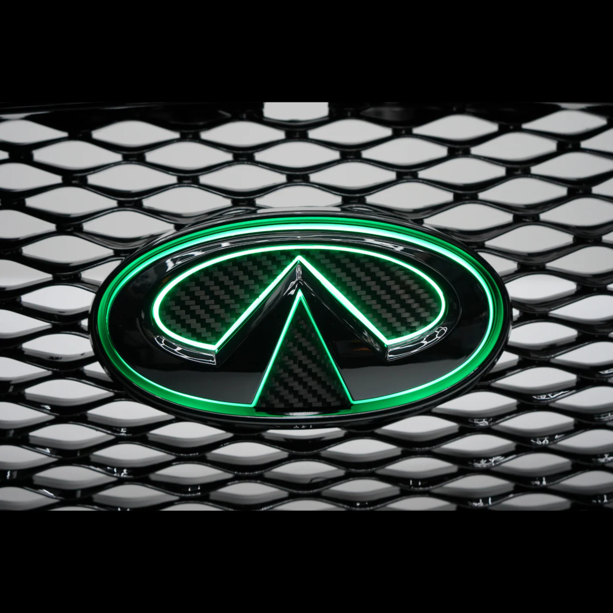 Close-up of the AutoJeweler gloss black and carbon fiber Infiniti Q50 LED grille emblem illuminated in bright green on the factory grille.