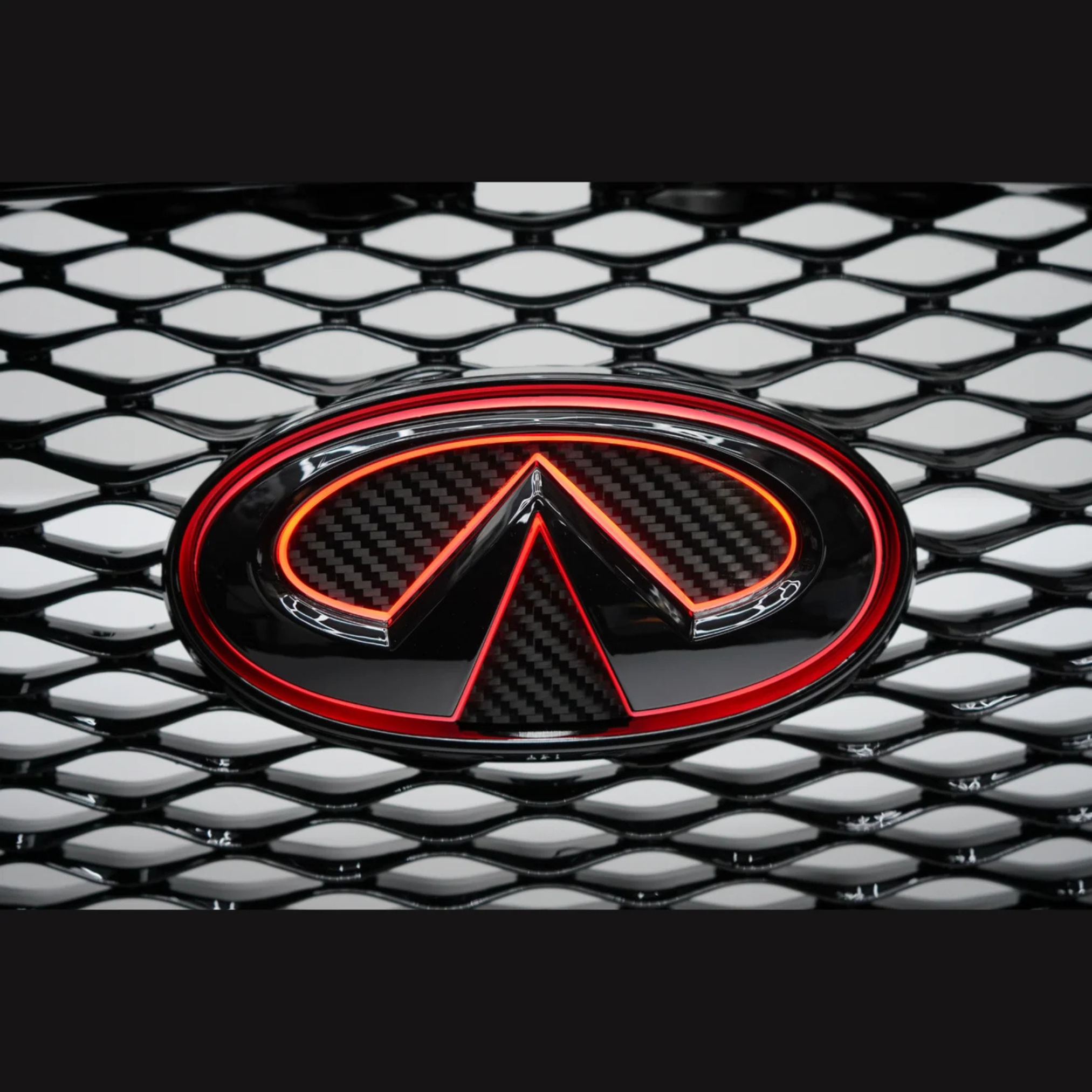 Close-up of a gloss black and carbon fiber Infiniti Q50 LED grille emblem illuminated in solid red on the factory mesh grille.