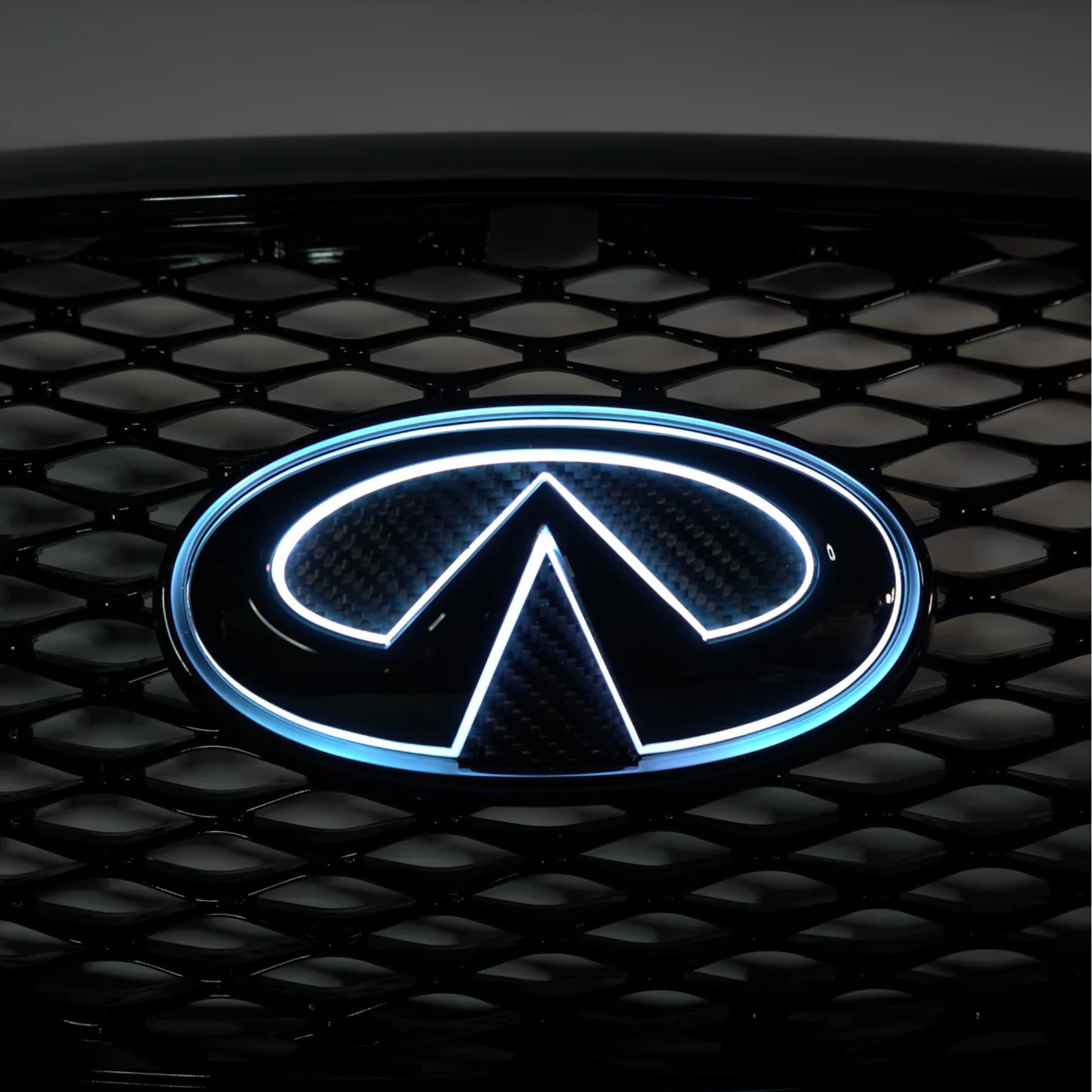 Close-up of a gloss black and carbon fiber Infiniti Q50 LED grille emblem illuminated on the factory mesh grille.