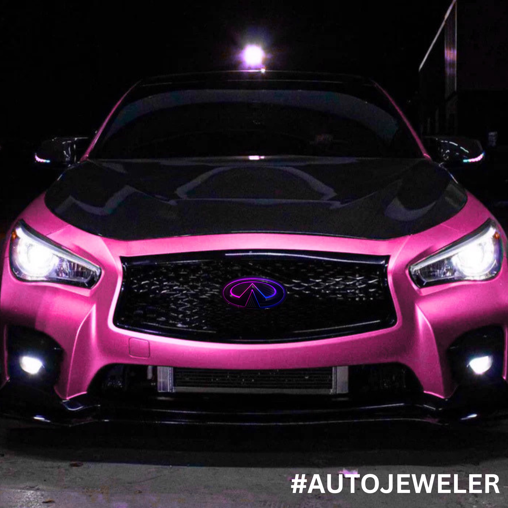 Front view of a pink wrapped Infiniti Q50 showcasing an AutoJeweler RGB Flow Series LED grille emblem illuminated with a purple and pink multicolor flow effect at night.