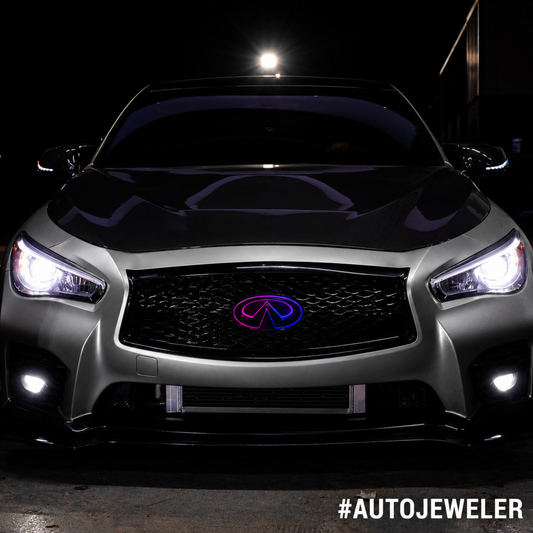 Front view of a silver Infiniti Q50 at night featuring an AutoJeweler RGB Flow Series LED grille emblem with a purple and blue multicolor flow effect installed.