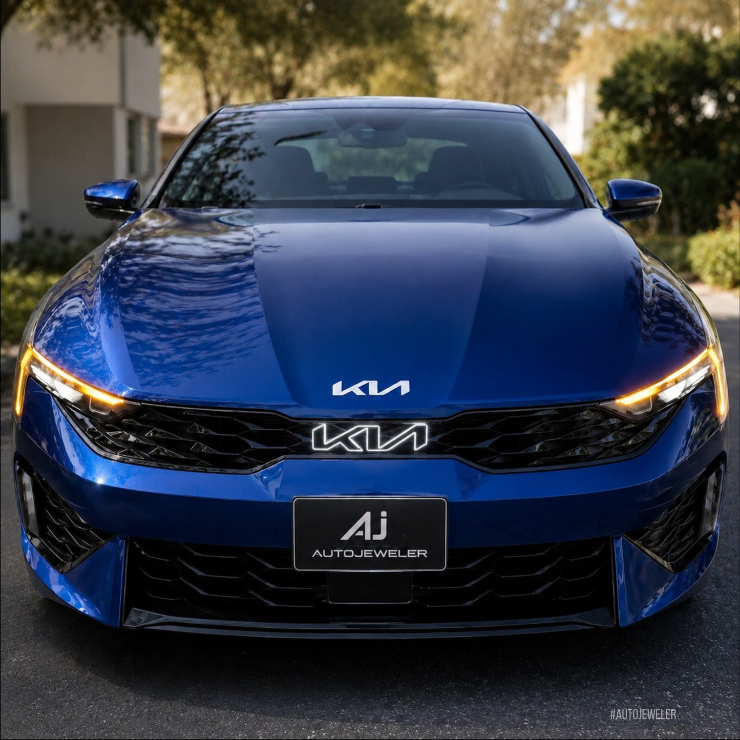 A front-facing view of a metallic blue Kia K5 sedan parked outdoors on a paved surface with trees and a building in the background. The OEM-style LED Kia logo has a white halo glowing around the logo making the emblem eye-catching, and a black AutoJeweler license plate frame mounted below.