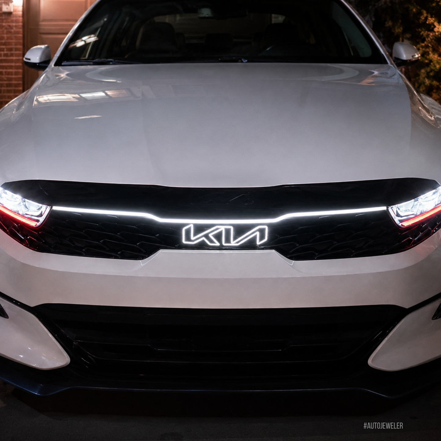A close-up front view of a white Kia sedan showing the illuminated LED light bar across the black grille and the lit Kia logo.