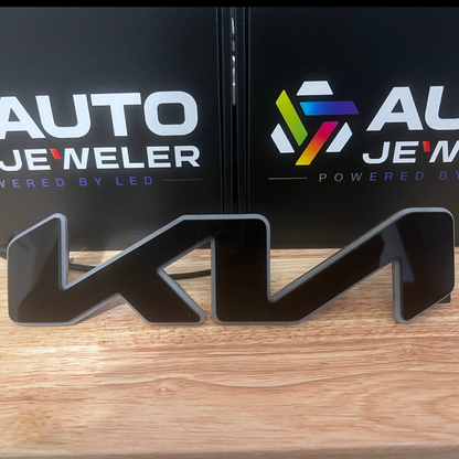A glossy black 3D LED light sign shaped like the letter 'K' on a wooden tabletop, with two branded black boxes behind it reading AUTO JEWELER.