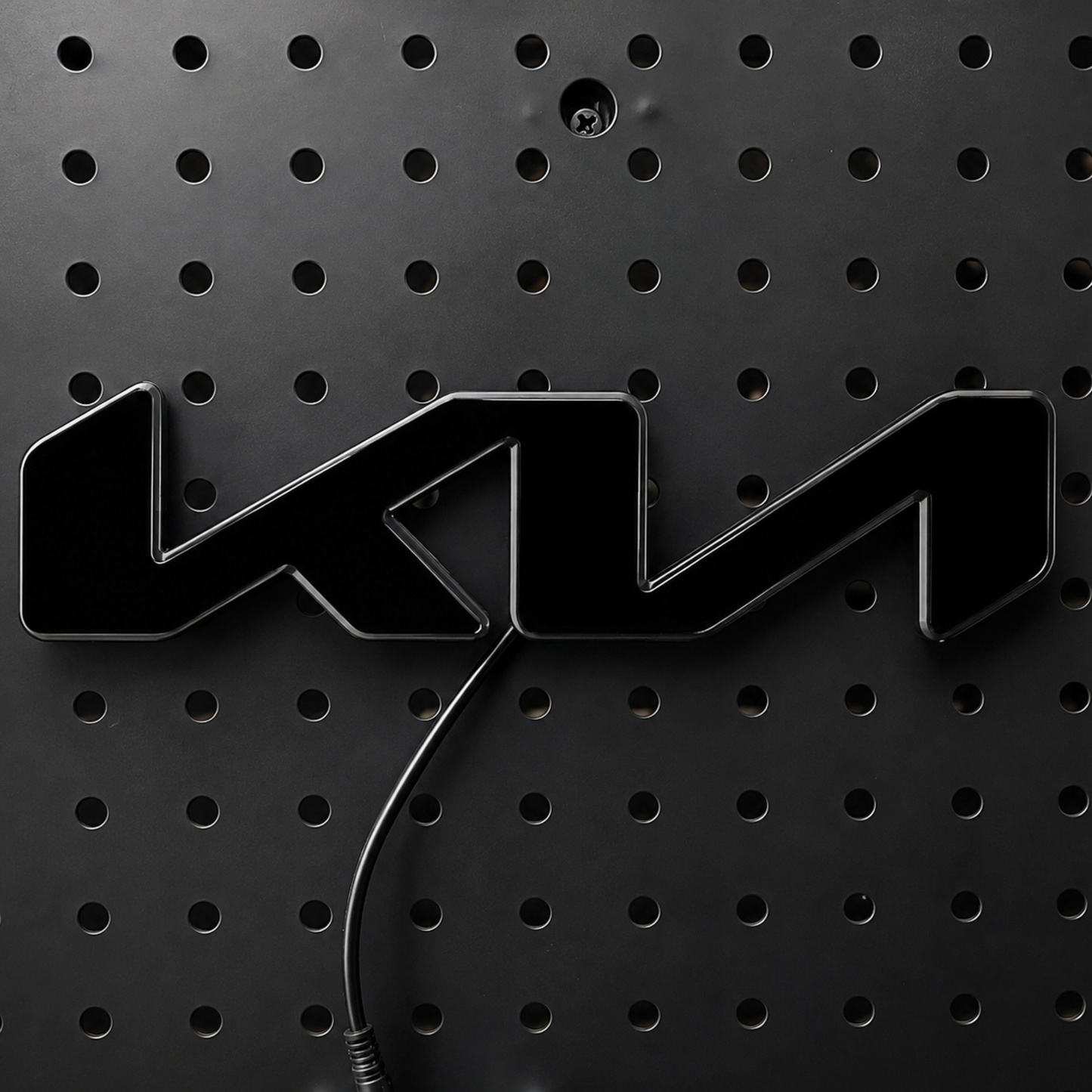 A glossy black 3D metal emblem featuring the stylized letters 'KM', mounted on a black perforated metal panel with a black cable visible below.