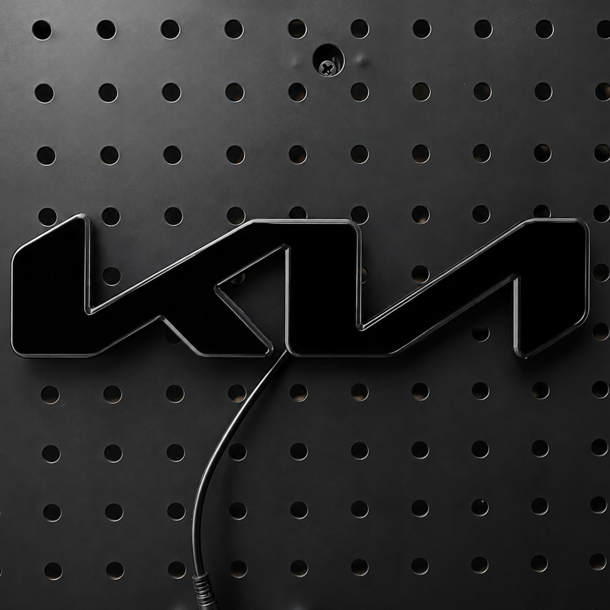 A glossy black 3D metal emblem featuring the stylized letters 'KM', mounted on a black perforated metal panel with a black cable visible below.