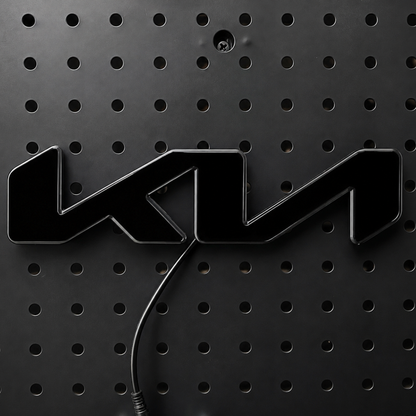 A glossy black 3D metal emblem featuring the stylized letters 'KM', mounted on a black perforated metal panel with a black cable visible below.