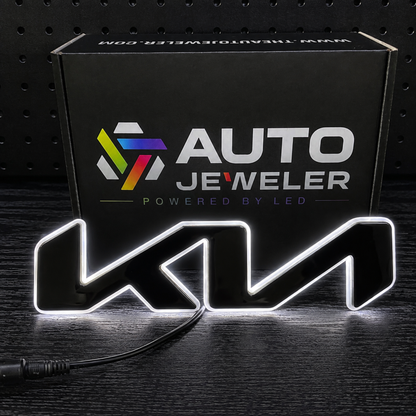 A black LED sign featuring the wordmark 'KIA' in a stylized font, illuminated with a white light border, shown in front of its black retail box branded 'AUTO JEWELER'.