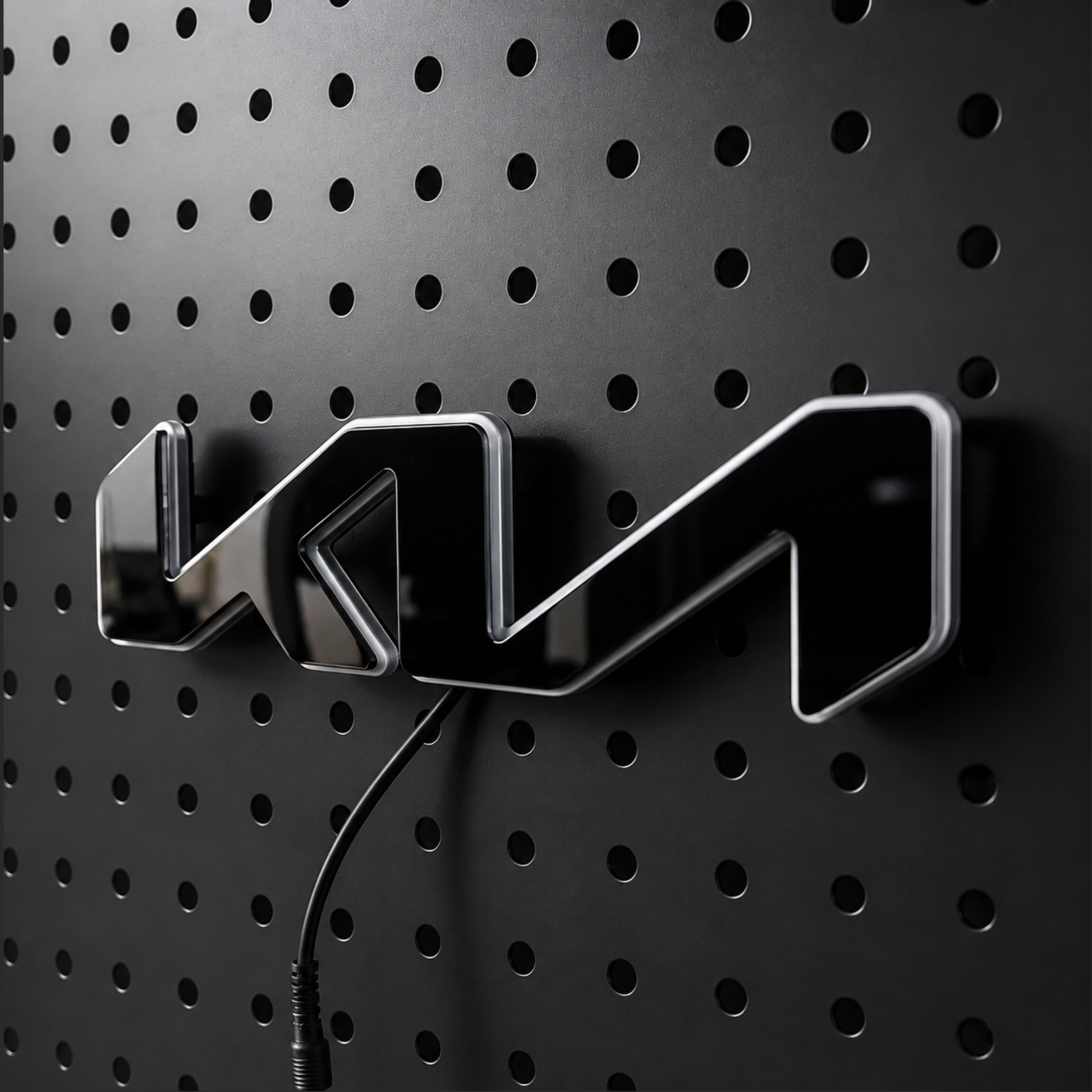 A glossy black 3D metallic emblem with silver edges, mounted on a black perforated metal wall. A black power cord is visible below the emblem.
