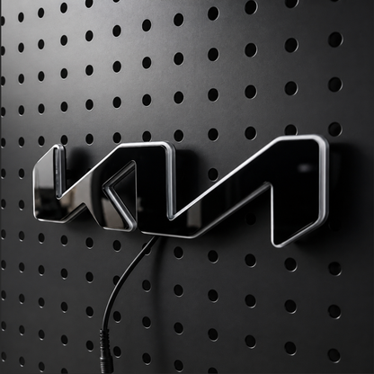 A glossy black 3D metallic emblem with silver edges, mounted on a black perforated metal wall. A black power cord is visible below the emblem.
