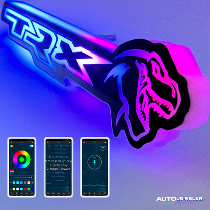 Close-up of illuminated TRX Flow Series grille emblem glowing blue and pink, with Bluetooth app control interface displayed on smartphones below and AutoJeweler branding in corner.