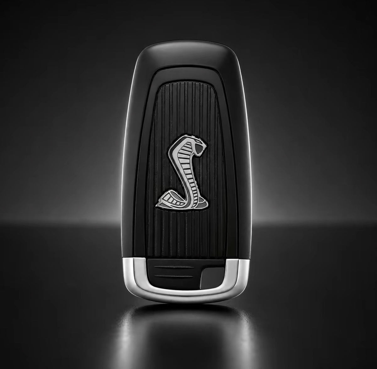 A black and silver car key fob featuring a silver cobra logo on a dark textured center, set against a dark gray gradient background.