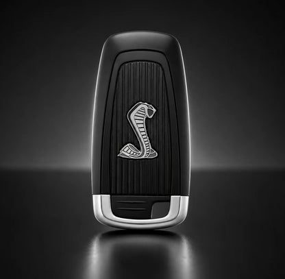 A black and silver car key fob featuring a silver cobra logo on a dark textured center, set against a dark gray gradient background.